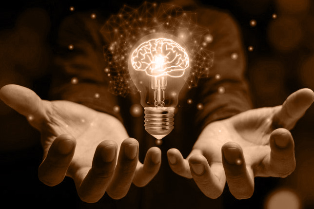 Sepia-toned image of a light bulb with the contours of a brain as the glowing filament, with a person holding their hands beneath it.